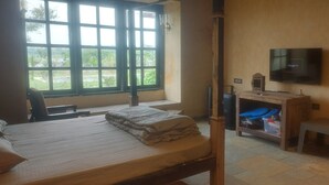 A room with a bed, a wooden desk, a television, and a window with a view of greenery. - Mura Yoga Villa (Kasrawad)