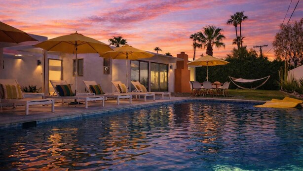 Un área junto a la piscina con sillones de descanso y sombrillas, una piscina y palmeras.