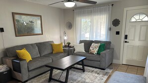 A living room with a gray sofa, a wooden coffee table, a gray ottoman, a painting on the wall, and a white door. - Peaceful Plum PA Home (Pittsburgh)