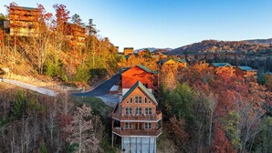 A large wooden cabin with a garage, surrounded by autumn foliage and a mountain backdrop. - Private Indoor Pool · Close 2 Dollywood · Mountain Views · Hot Tub + Game Room (Sevierville)