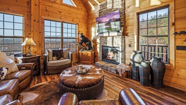 A cozy cabin interior with a fireplace, leather furniture, and large windows offering scenic views. - Private Indoor Pool · Close 2 Dollywood · Mountain Views · Hot Tub + Game Room (Sevierville)