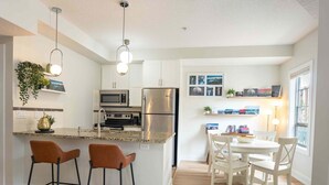 A modern kitchen with a central island, bar stools, a dining table with chairs, and a refrigerator. - 13th Ave Condo + City Views + Balcony + Walkable (Calgary)