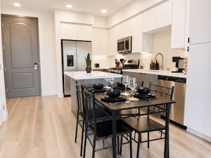 Una cocina moderna con electrodomésticos de acero inoxidable, un comedor con sillas negras y una mesa preparada para cuatro, y una puerta gris.