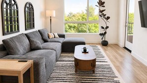 A modern living room with a sectional sofa, a wooden coffee table, a floor lamp, and a large window with a view of greenery. - Modern Apartment w/ Pool, Gym & Near Disneyland (Santa Ana)