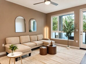 A modern living room with a beige sectional sofa, two round wooden side tables, a floor lamp, and a large window with a view of greenery. - Modern Apartment 10 Min to Disneyland | Pool & Gym (Santa Ana)