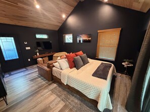 A bedroom with a bed, a desk, a television, and a window with blinds.