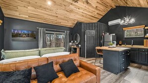 A modern interior with a wooden ceiling, a cozy living area with a leather sofa, a kitchen with dark cabinetry, and a large window offering a view of the outdoors. - Honeymoon Hideaway • Hot Tub • Fire Pit (Sevierville)