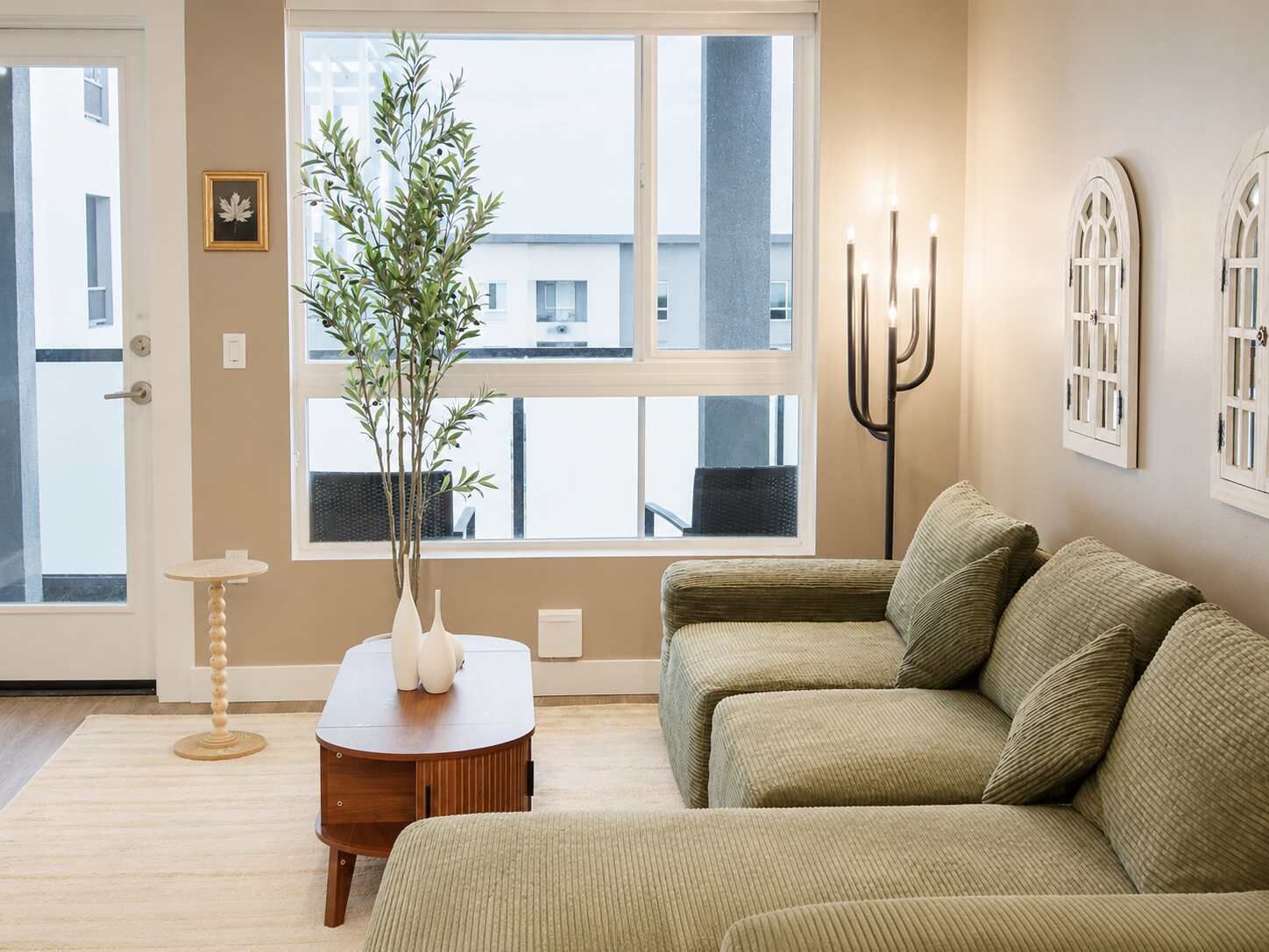 A living room with a sofa, a small table with a plant, a floor lamp, and a window with a view of buildings.