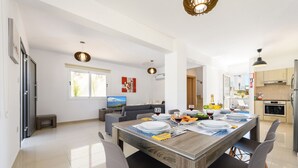 A modern kitchen with a dining area, featuring a wooden table set for four, a sofa, and a TV in the background. - Nissini Villa 8 in Ayia Napa (Ayia Napa)
