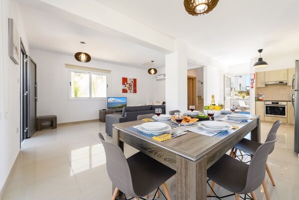 A modern kitchen with a dining area, featuring a wooden table set for four, a sofa, and a TV in the background.