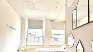 Een moderne slaapkamer met een groot raam, een bed met een beige deken, een bruine bank en meerdere spiegels aan de muur.
