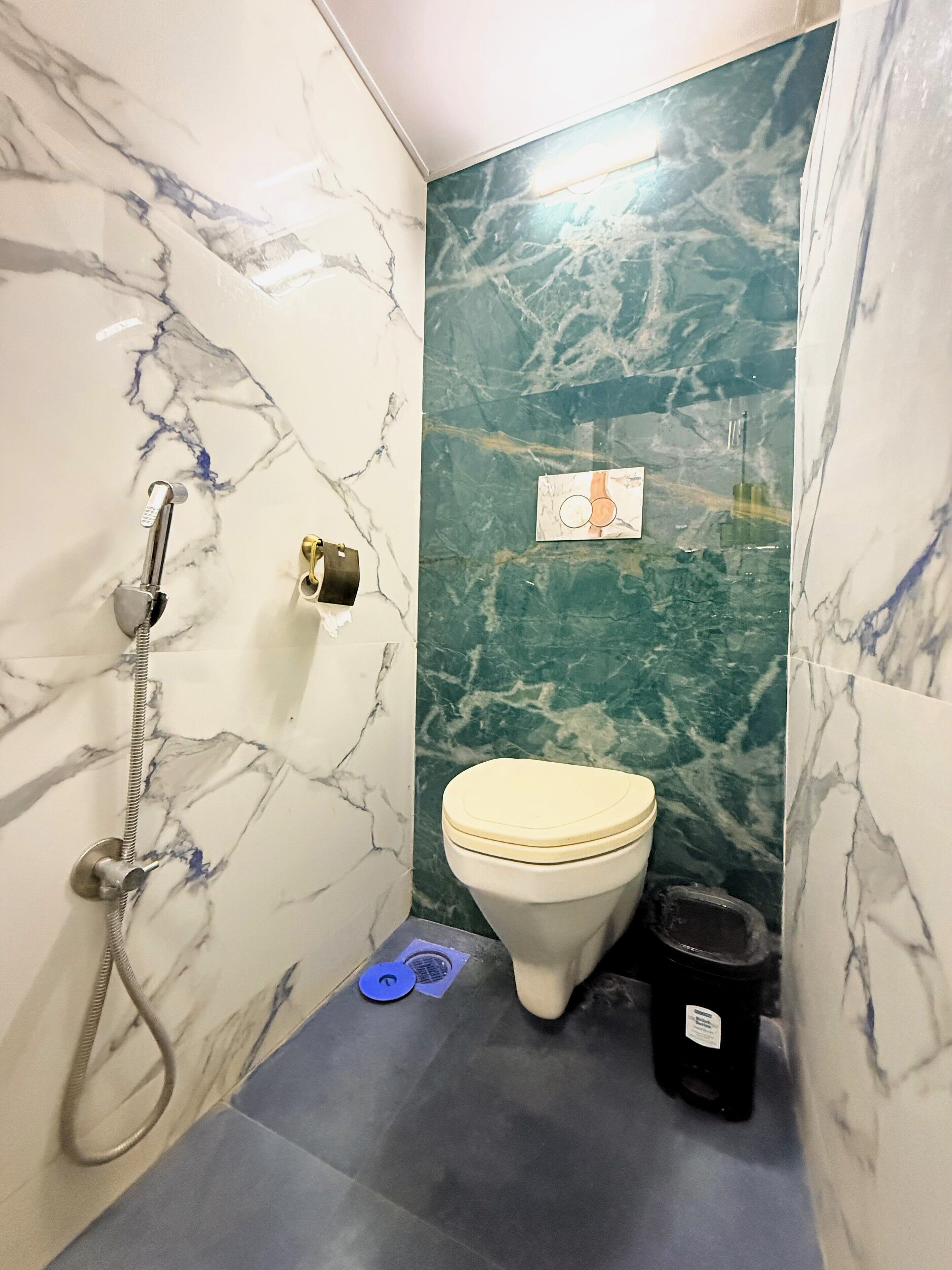 A bathroom with marble walls, a toilet, a shower, and a trash bin.
