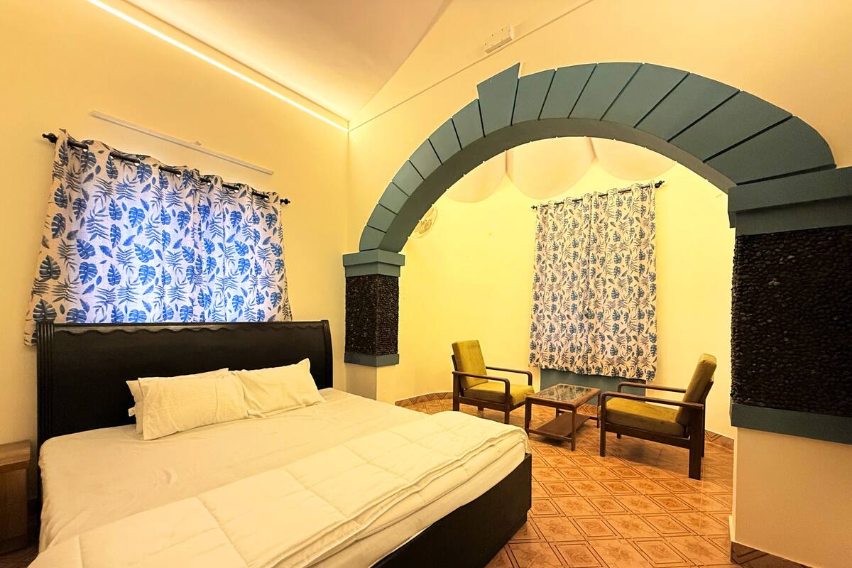 Habitación de hotel con una cama grande, dos sillas, una mesita pequeña y un arco decorativo.