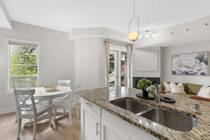 A modern kitchen with a granite countertop, double sink, and white cabinetry. There is a dining area with a round table and chairs, and a large window providing natural light. - 13th Ave - DT Calgary - 2BD 2BA - Sleeps 5 (Calgary)