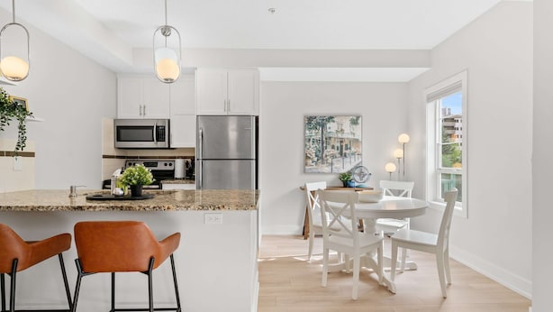 A modern kitchen with white cabinets, stainless steel appliances, and a granite countertop. There are two bar stools and a dining table with white chairs. - 13th Ave - DT Calgary - 2BD 2BA - Sleeps 5 (Calgary)