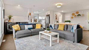 A modern living room with a grey sectional sofa, a white coffee table, and a patterned rug. A washing machine is visible in the background, indicating a laundry area. - Local Loft Downtown Okotoks 2BD Private (Okotoks)