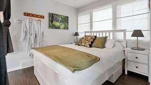 A neatly made bed with a white headboard and a green throw blanket, a bedside table with a lamp, and a window with white curtains. - Local Loft Downtown Okotoks 2BD Private (Okotoks)