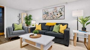 A modern living room with a grey sofa, a white coffee table, and a black sofa with yellow cushions. There is a painting on the wall, a lamp, and a potted plant. - The Owlet Nwcalgary Queenbd Privatesuite (Calgary)