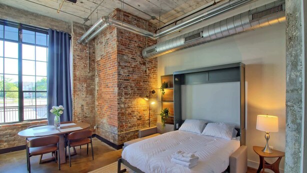 Una habitación de hotel moderna con pared de ladrillo, una cama grande, un escritorio con silla y una ventana con cortinas.
