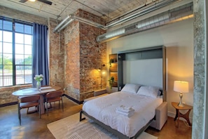 Una habitación de hotel moderna con pared de ladrillo, una cama grande, un escritorio con silla y una ventana con cortinas.