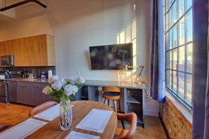 A modern kitchen with wooden cabinets, a dining table with papers, and a vase of white flowers. - Chic Ultramodern Studio Loft w/ Designer Features (Saint Louis)