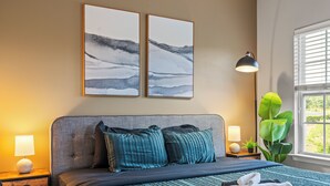 A neatly made bed with a striped bedspread, two pillows, a bedside lamp, and a nightstand with a lamp. There are two framed pictures on the wall above the bed. - Upscale King 3BR Townhouse in Pineville (Pineville)