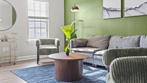 A modern living room with a round wooden coffee table, a green sofa, and a grey sofa. There is a window with blinds, a green wall, and a floor lamp. - Upscale King 3BR Townhouse in Pineville (Pineville)