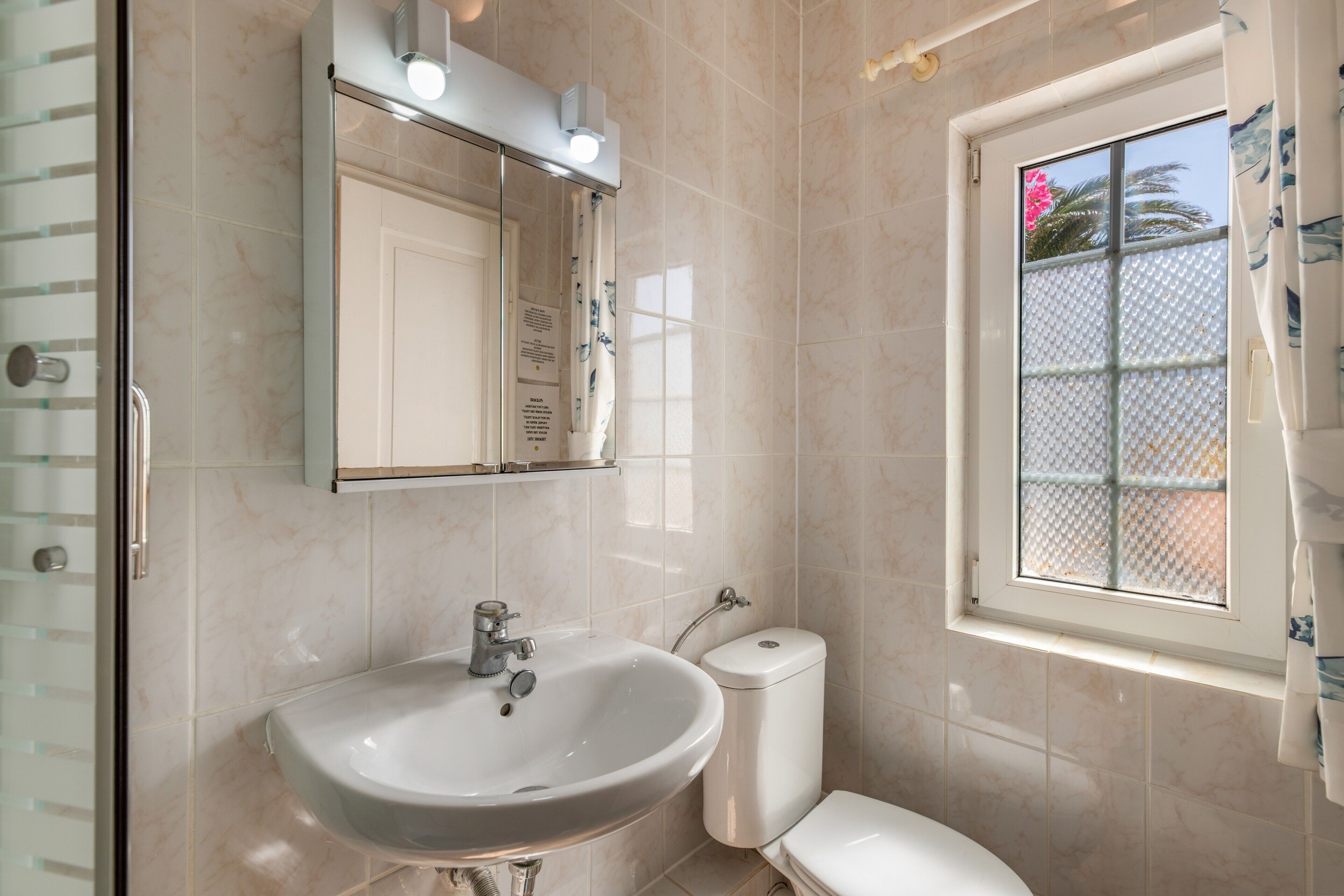 Un baño con lavamanos, inodoro y ventana con vista a palmeras.