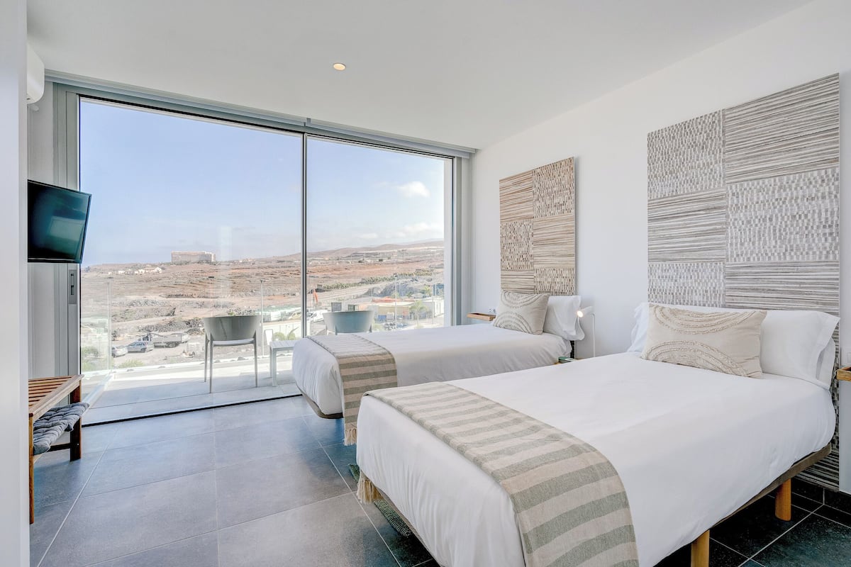 A modern hotel room with two beds, a large window with a city view, and a flat-screen TV on the left.
