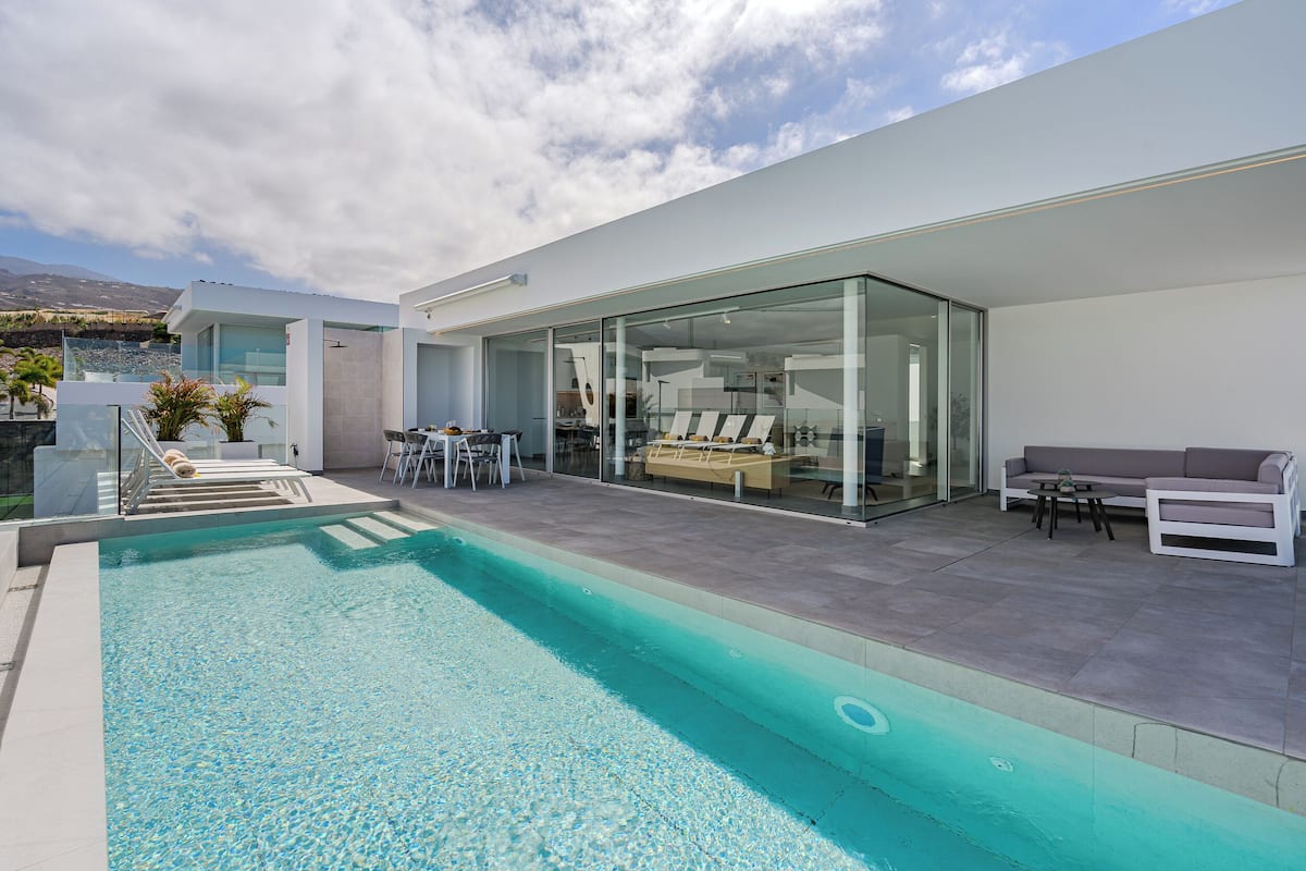 A modern outdoor pool area with a glass-enclosed lounge, a seating area with a sofa and table, and a staircase leading to an upper level.