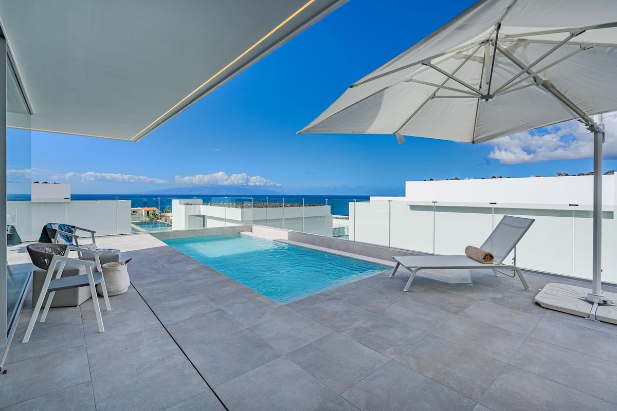 A rooftop pool area with a clear view of the ocean, a white umbrella, and modern lounge chairs.