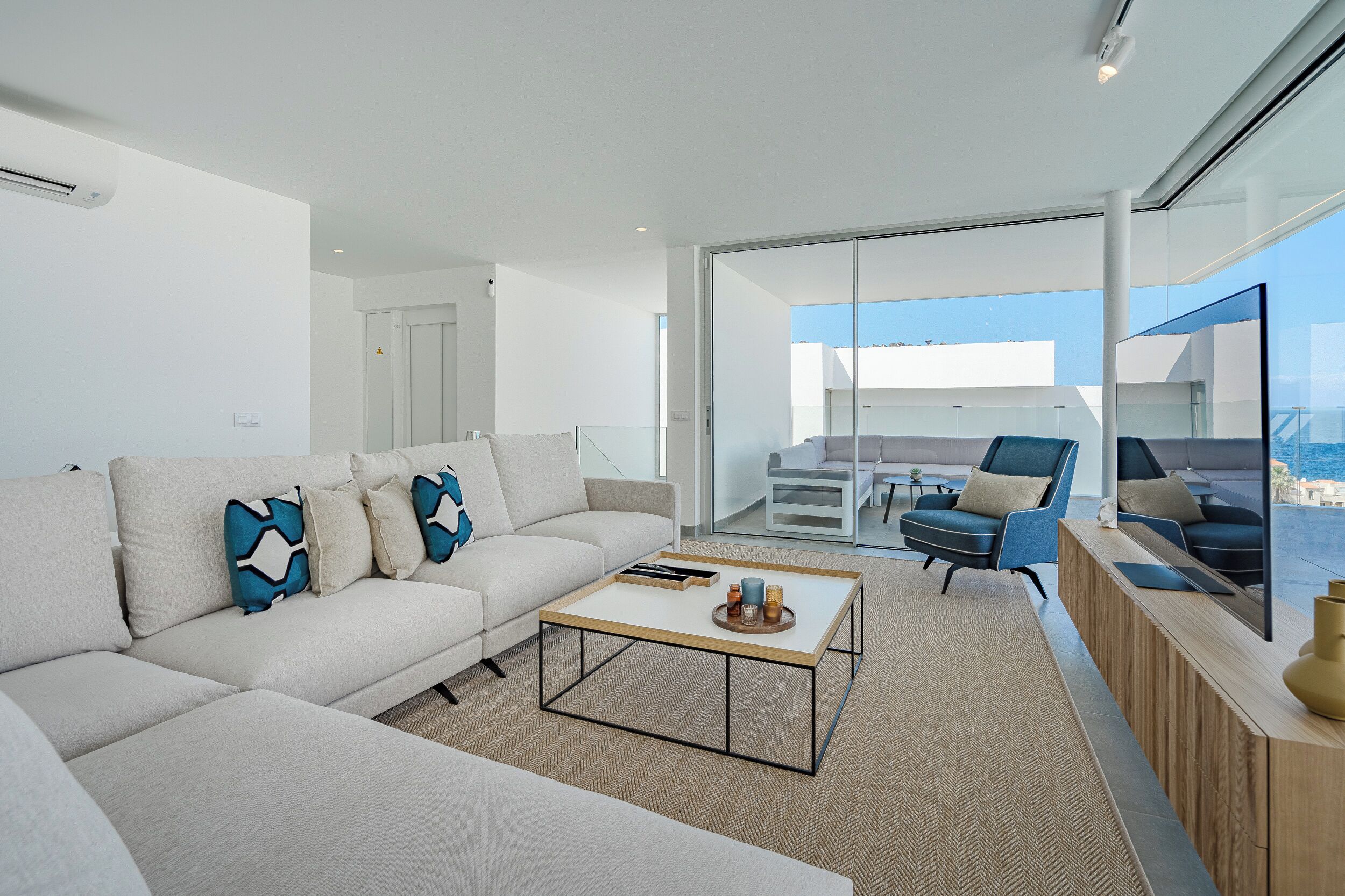 A modern living room with a sofa, coffee table, and chairs. There is a glass door leading to a balcony with a view of the sea.