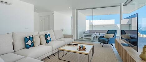A modern living room with a sofa, coffee table, and chairs. There is a glass door leading to a balcony with a view of the sea.