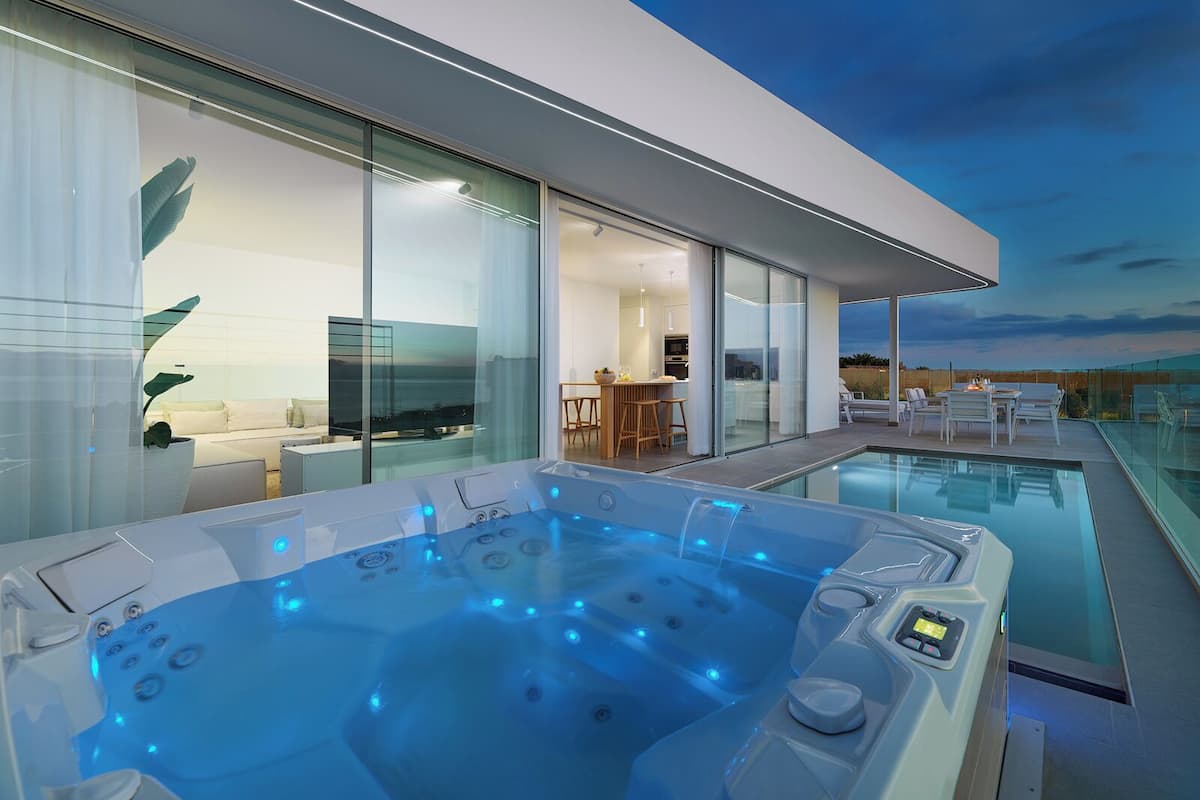 A modern outdoor hot tub area with a glass-enclosed pool, a dining area, and a view of the sunset.