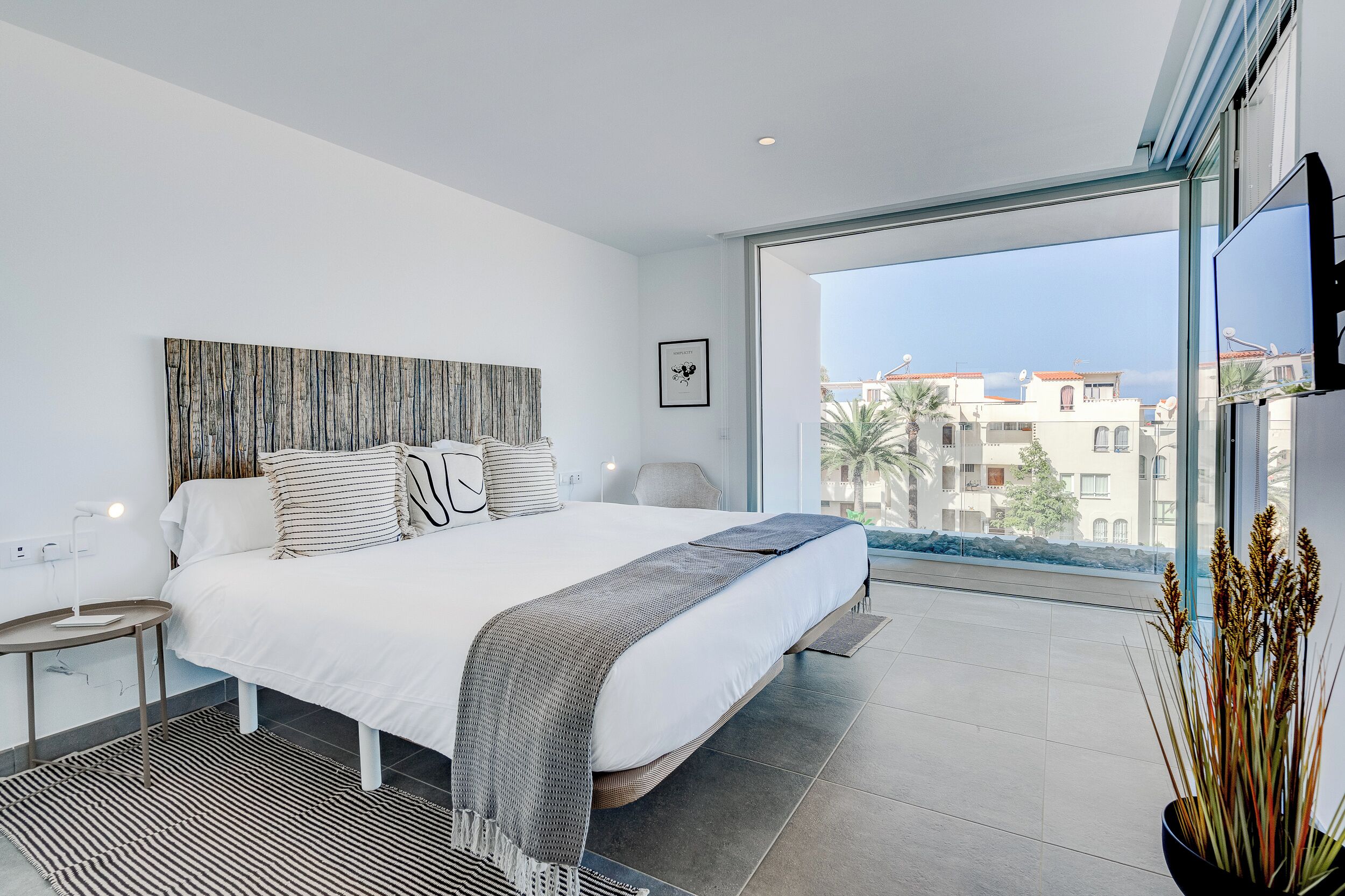 Ein modernes Schlafzimmer mit einem großen Bett, Blick auf Gebäude durch ein großes Fenster und einem Blumentopf auf der Fensterbank.