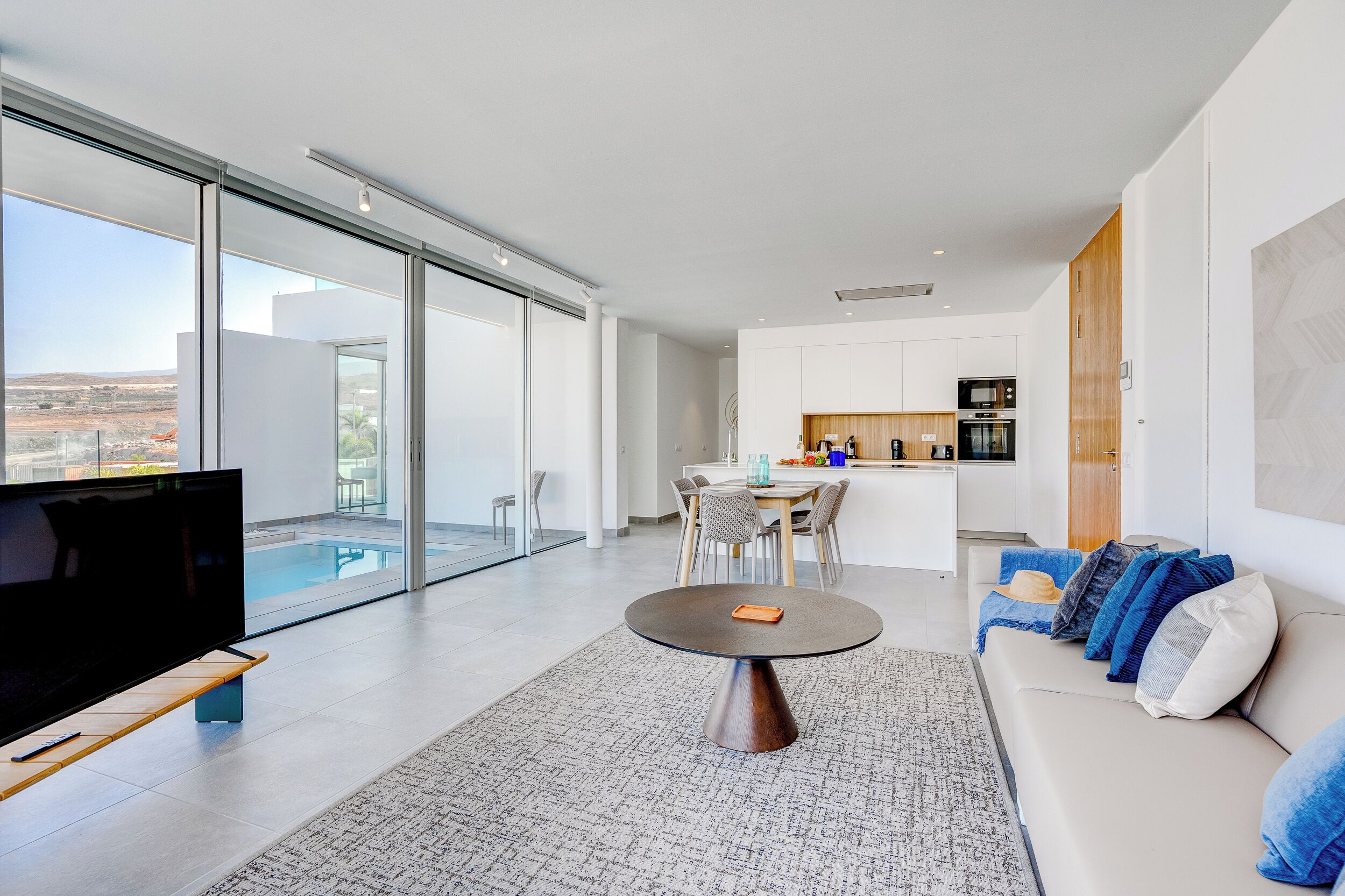 A modern living room with a large TV, a round coffee table, and a sofa with blue and white cushions. There is a dining area with a table and chairs, and a kitchen in the background.
