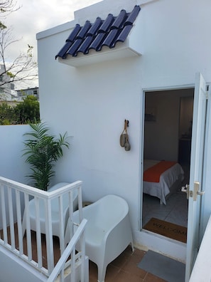 Un edificio blanco con techo de tejas, una puerta blanca y un felpudo con la palabra 'WELCOME', una pequeña silla blanca, una planta en maceta y una barandilla blanca.