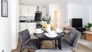 A modern kitchen with white cabinets, a black stove, and a stainless steel range hood. There is a dining area with a black table and grey chairs, and a television in the living room. - Chic 1BR Close to Buckingham Palace (London)