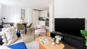 A modern living room with a large flat-screen TV, a coffee table with magazines, a sofa, and a dining area with a chair and a small table. - Chic 1BR Close to Buckingham Palace (London)