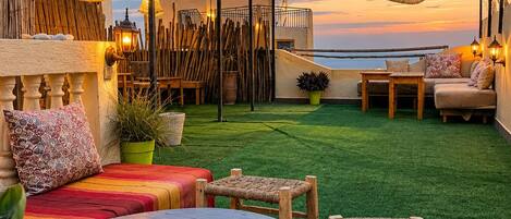 An outdoor seating area with a round table, chairs, and a sofa, overlooking a body of water at sunset.