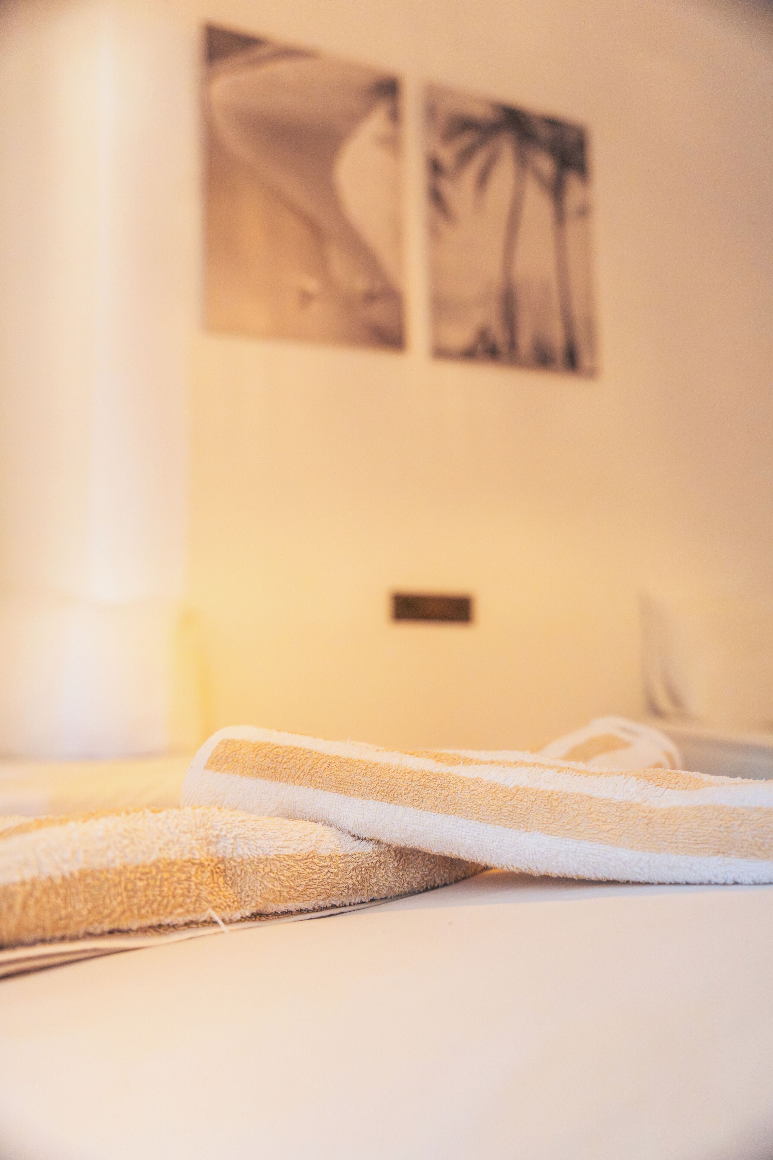 Una cama con una toalla blanca y color crema, una mesita de noche y dos cuadros en la pared que muestran una mujer y palmeras.