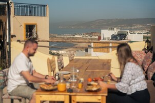 Una mesa de comedor con comida y bebidas, vista al mar y edificios al fondo.