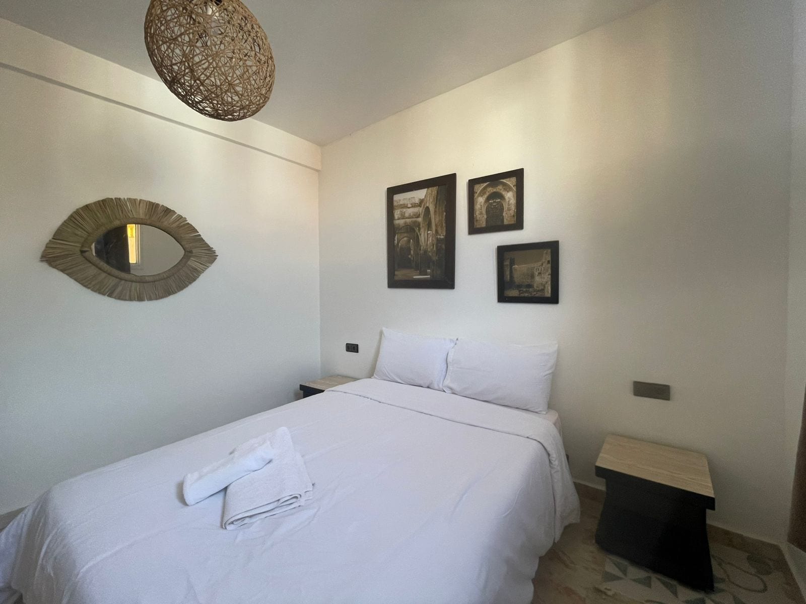 A neatly made bed with white linens and a folded towel, a round mirror on the wall, and three framed pictures above the bed.