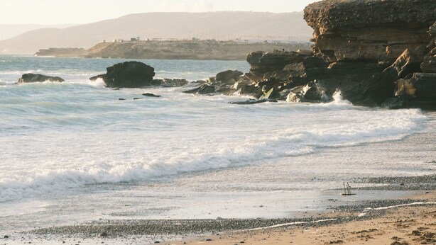 A coastal beach with waves, a sailboat, and rocky cliffs. - Eco Bay Surf Camp (Aourir)