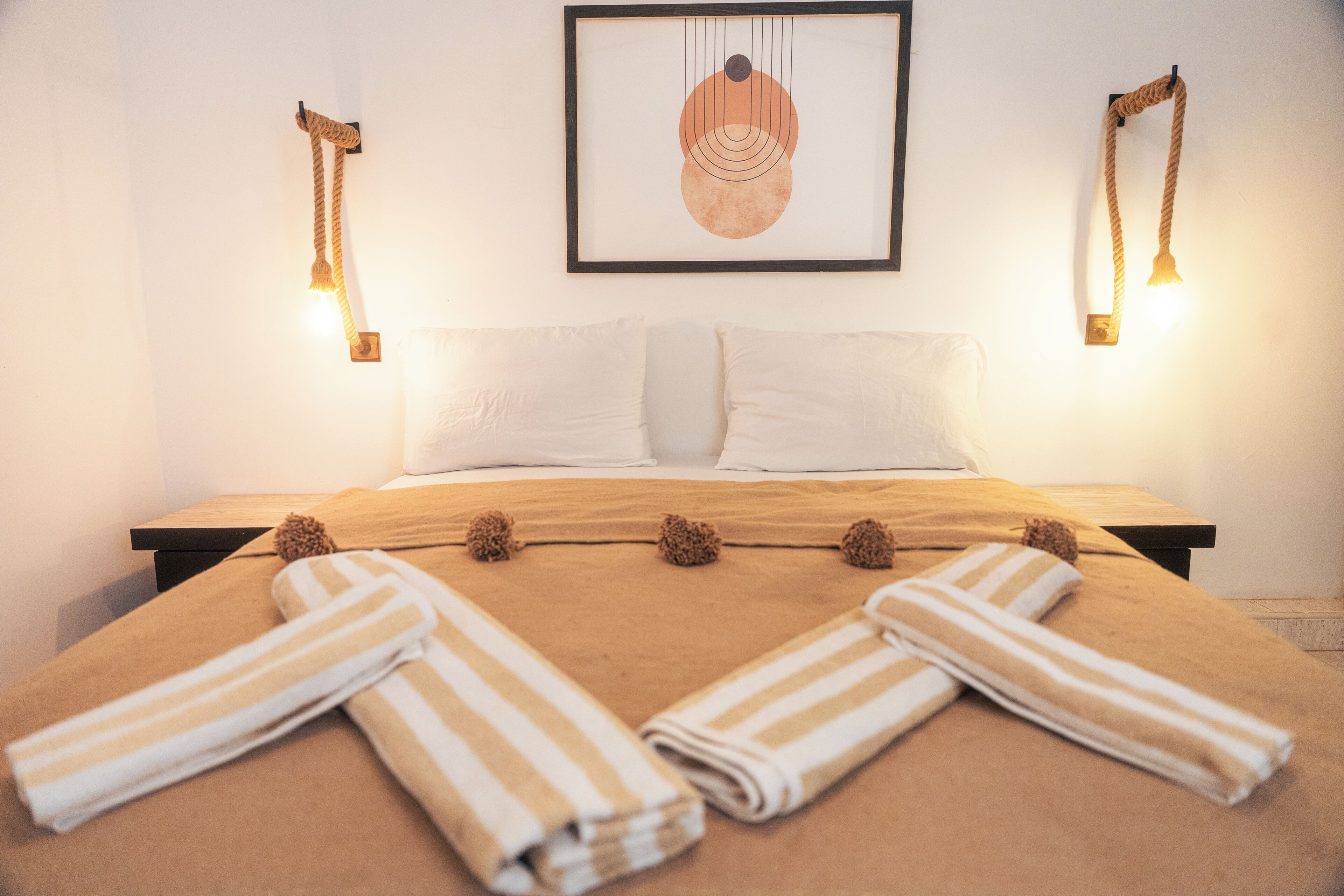 A neatly made bed with striped towels, a framed artwork above, and two wall-mounted lamps on either side.