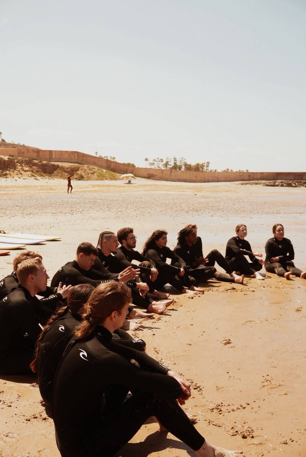 A group of people in wetsuits sitting on a sandy beach.