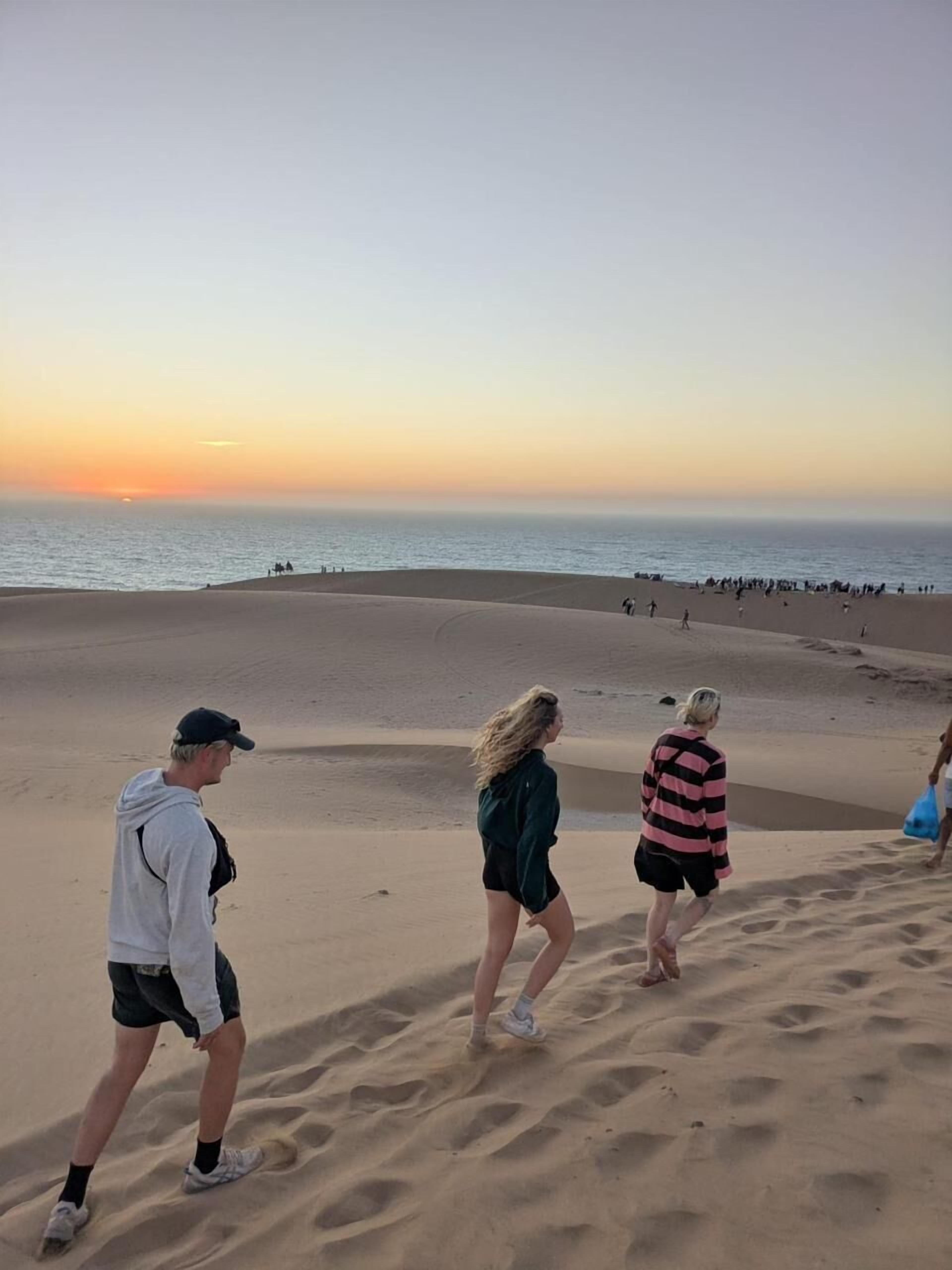 Cena de praia com pessoas caminhando na areia ao pôr do sol.