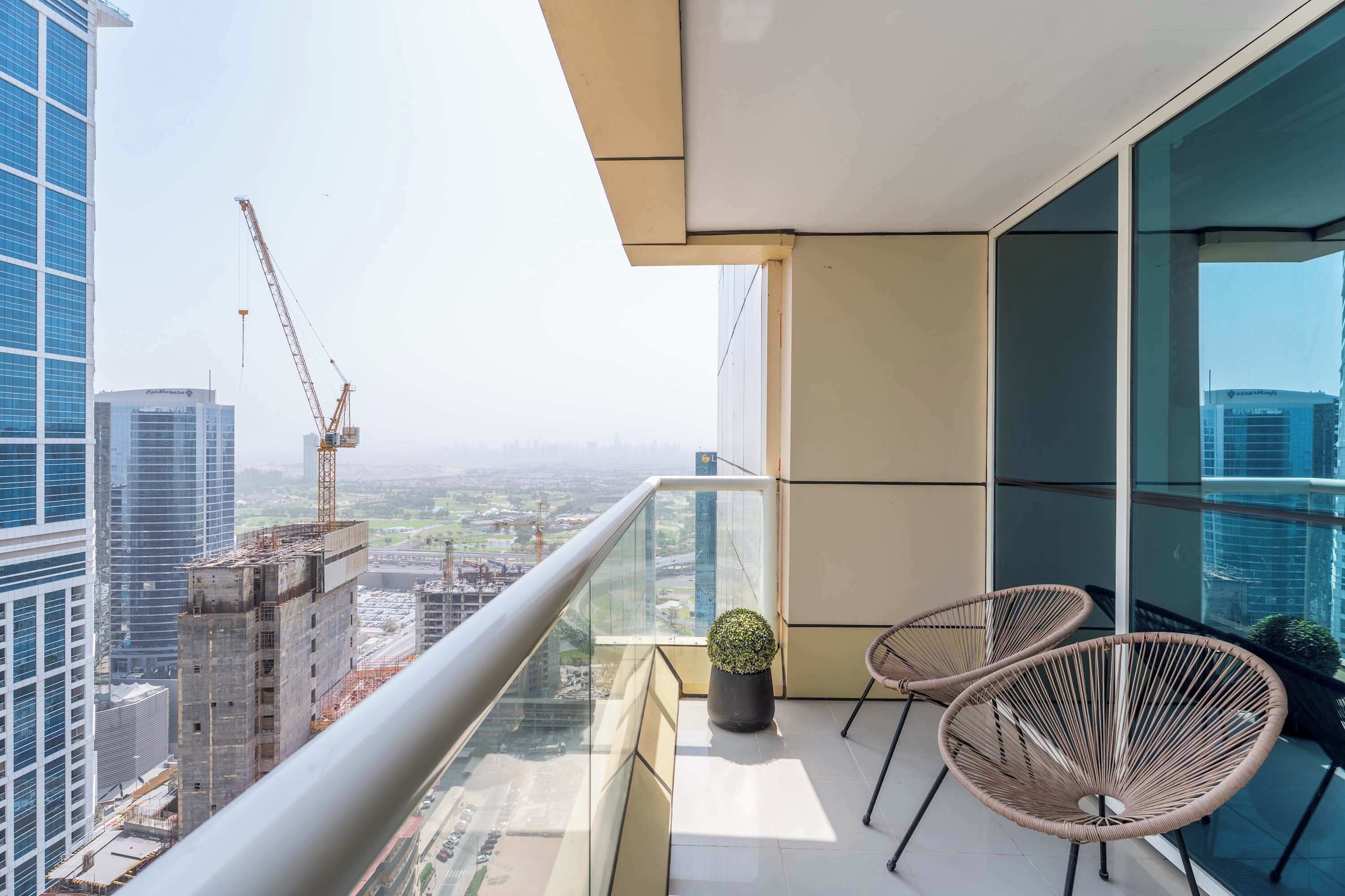 A balcony with a view of a cityscape, including construction cranes, and two wicker chairs with a potted plant.