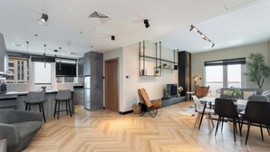 Een ruime woonkamer met een visgraatvloer, een eethoek met een tafel en stoelen, een keuken met moderne apparatuur, en een groot raam dat zorgt voor natuurlijk licht.