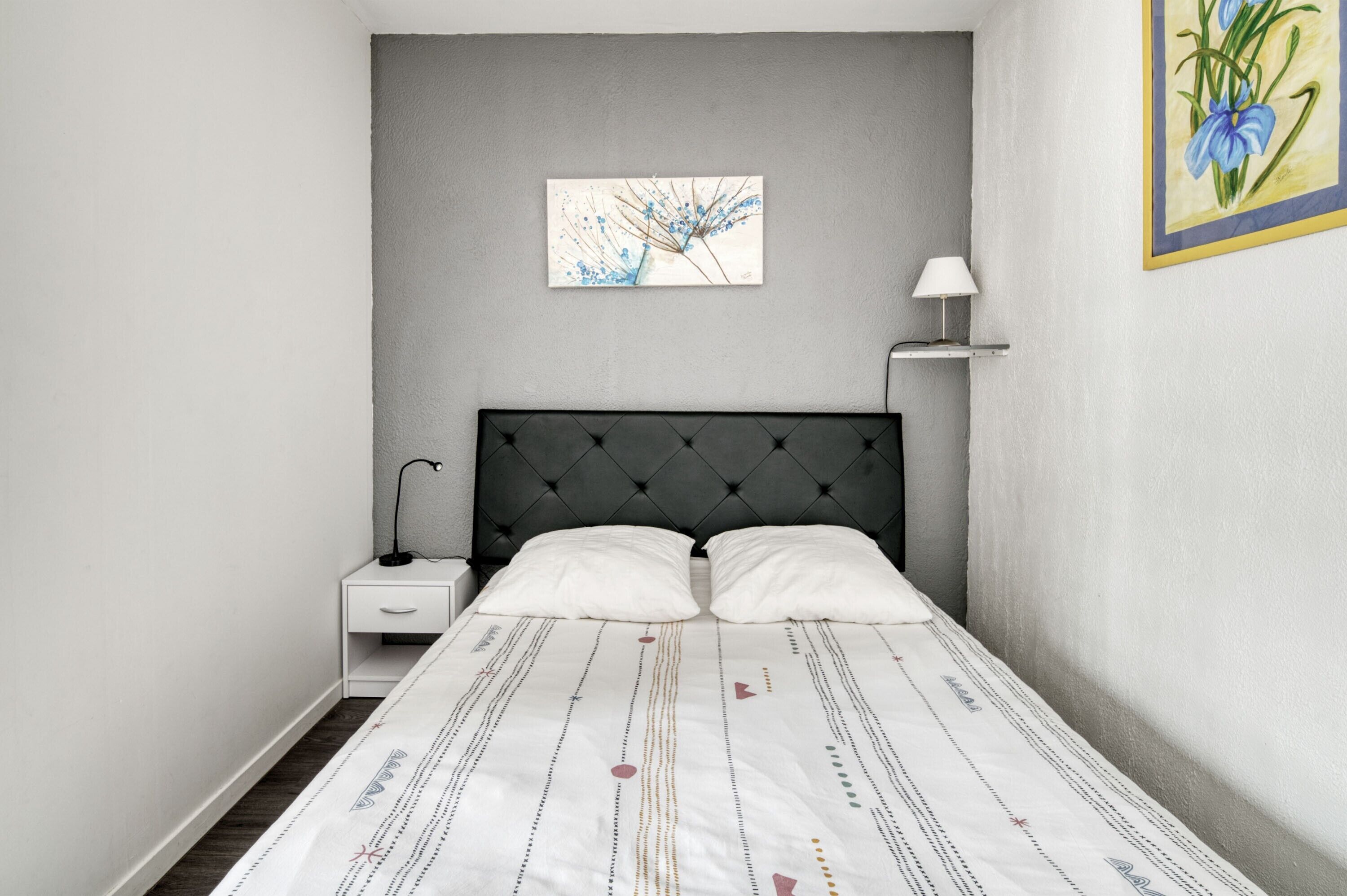 Un lit bien fait, recouvert d’une couvre-lit à motifs, avec une tête de lit noire à capitons, une table de chevet dotée d’une lampe et une œuvre d’art accrochée au mur.
